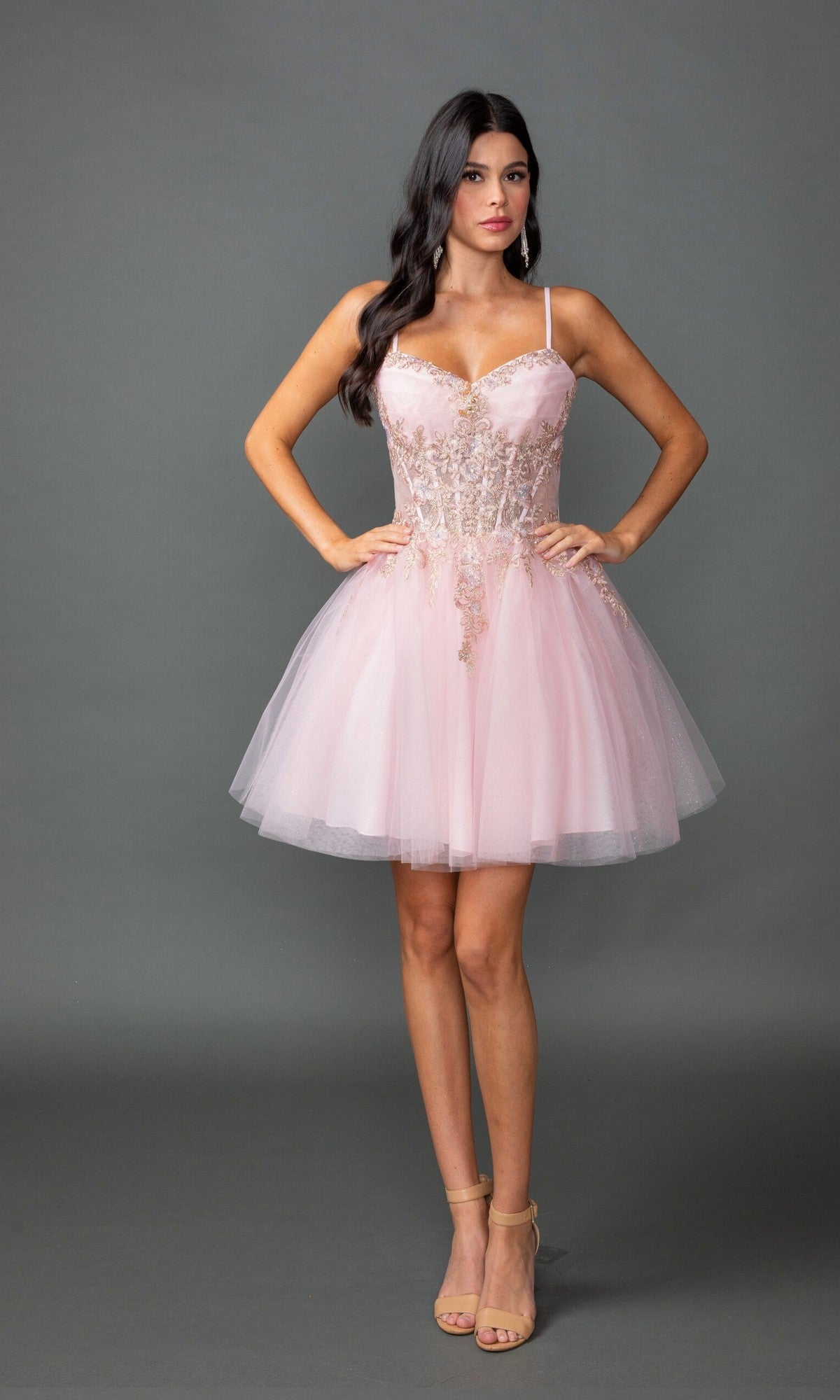 A woman poses against a gray background in the Short Prom Dress: Dancing Queen 3424—a light pink dress with a sheer corset bodice, beaded lace embroidery, and spaghetti straps—styled elegantly with nude heels.